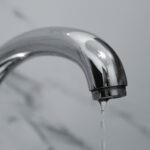 A close-up shot of a modern chrome bathroom faucet with water dripping from the tip.