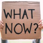 Cardboard sign reading 'What Now?' held outdoors, conveying uncertainty or protest.